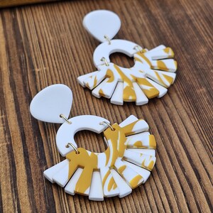 May include: A pair of white and yellow earrings. Each earring features a white teardrop-shaped top connected to a white semi-circle with a yellow and white fan-shaped design. The earrings are displayed on a wooden surface.