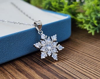 May include: A silver snowflake pendant necklace with clear crystals. The pendant is a detailed snowflake design with a large central crystal and smaller crystals on each point. The necklace is displayed on a blue surface.