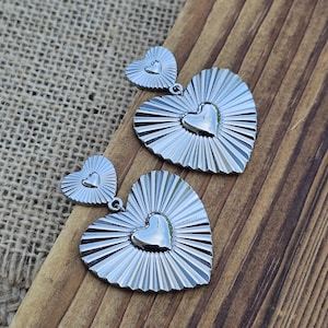May include: Silver heart-shaped earrings. Each earring has a large heart with a textured, sunburst design and a smaller heart accent. The earrings are displayed on a wooden surface.