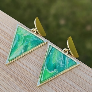 May include: A pair of gold-coloured earrings with a triangular design. The earrings have a marbled green and white pattern within a gold frame, with a small gold accent at the top. The earrings are displayed on a wooden surface.