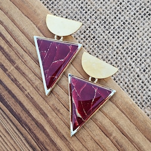 Op de afbeelding: Een paar goudkleurige oorbellen. Elke oorbel heeft een gouden halve cirkel stud en een driehoekige hanger met een zilveren rand. De hanger is gevuld met dieprode hars met gouden aderen. De oorbellen worden gepresenteerd op een houten oppervlak.