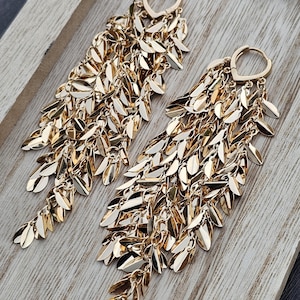Puede incluir: Un par de pendientes colgantes dorados. Cada pendiente presenta un cierre de aro y múltiples hilos de pequeños abalorios en forma de hoja. Los pendientes se exhiben sobre una superficie de madera clara.