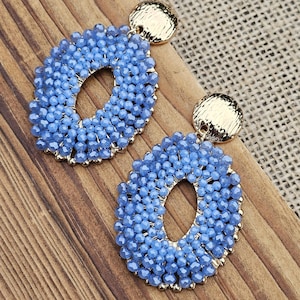 May include: A pair of blue beaded oval earrings with gold-colored circular studs. The earrings are made of small, light blue beads arranged around an oval shape. The earrings are displayed on a wooden surface.
