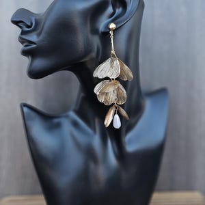 Puede incluir: Pendientes colgantes de tono dorado con un diseño de hojas superpuestas y una cuenta blanca en forma de lágrima en la parte inferior.