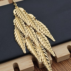 Puede incluir: Pendientes dorados con forma de hojas. Estos pendientes presentan múltiples colgantes en forma de hojas superpuestas, creando un efecto en cascada. Están hechos de un material metálico brillante y están diseñados para colgar. El fondo es oscuro con detalles de madera.