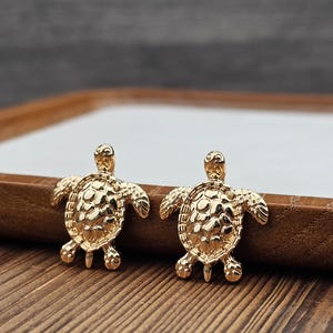 May include: A pair of gold-tone turtle stud earrings. The turtles are detailed with a textured shell and have a three-dimensional design.