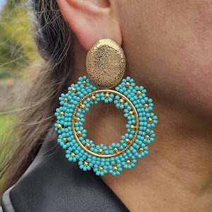 May include: Gold-coloured stud earrings with a textured surface and a turquoise beaded hoop design. The hoop is adorned with small gold-coloured beads. These earrings are a statement piece.