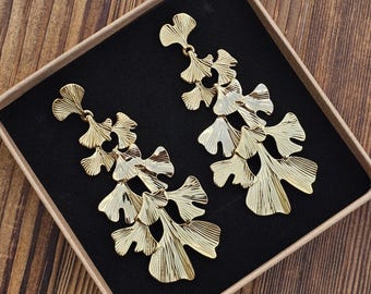 May include: A pair of gold-colored leaf-shaped earrings displayed in a black box. The earrings have a layered design with multiple leaf-like elements. The box is set against a wooden background.