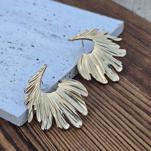 May include: A pair of gold-colored, fan-shaped earrings. The earrings have a textured, layered design, resembling stylized leaves or feathers. The earrings are displayed on a light-colored stone surface against a wooden background.