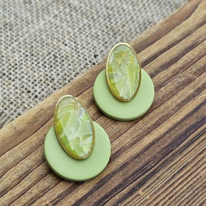 May include: A pair of handmade earrings. Each earring features a light green circular base with a gold-toned oval top. The oval tops have a marbled green and yellow design. The earrings are displayed on a wooden surface.