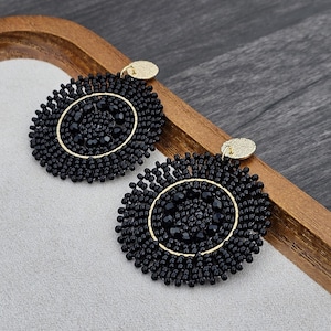 Puede incluir: Un par de pendientes redondos de cuentas negras con detalles dorados. Los pendientes son redondos y tienen un diseño circular en el centro.
