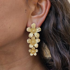 May include: Gold flower dangle earrings with three flowers cascading down. The flowers are textured and have a delicate design.