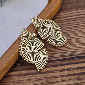May include: A pair of gold-tone earrings with a geometric fan design. The earrings are made of metal and have a delicate, intricate pattern.