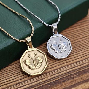 Puede incluir: Dos collares con colgantes de mariposa. Uno es dorado, el otro plateado. Cada colgante tiene forma de octágono con un diseño de mariposa. Los collares están sobre una superficie de madera con una caja verde en el fondo.