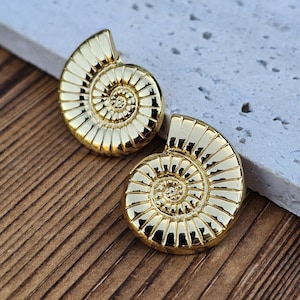 May include: A pair of gold-coloured earrings, each shaped like a spiral. The earrings have a detailed, ridged pattern. They are displayed on a light-coloured, textured surface, with a wooden background.