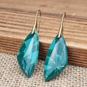 Puede incluir: Un par de pendientes colgantes de color turquesa y dorado. Los pendientes tienen forma de lágrima con un patrón de mosaico de turquesa y dorado. Los ganchos son de color dorado.