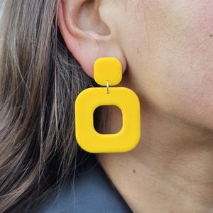 Puede incluir: Un par de pendientes geométricos amarillos con forma cuadrada y un recorte cuadrado más pequeño en el centro. Los pendientes están hechos de un material liso y mate.