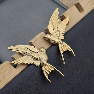Pode incluir: Dois objetos decorativos dourados em forma de pássaro com asas abertas. Os pássaros são detalhados com penas texturizadas e longas penas da cauda. Eles estão em uma superfície escura com um suporte de madeira.