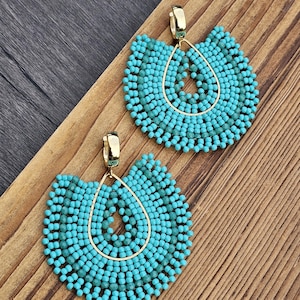 May include: A pair of turquoise beaded earrings with gold-colored accents. The earrings have a teardrop shape with a gold frame and are made of small, round beads. The earrings are displayed on a wooden surface.