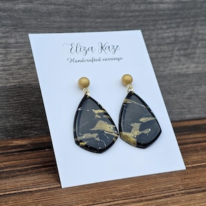May include: Black and gold dangle earrings with a marbled design. The earrings are on a white card with the text "Eliza Kaze Handcrafted earrings".