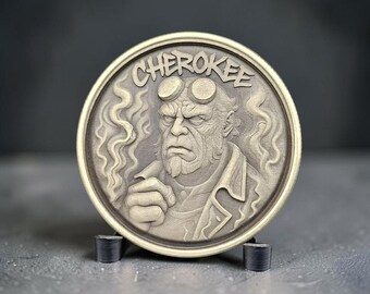 Puede incluir: Una moneda metálica circular con un retrato en relieve de un personaje con cuernos y perilla, vistiendo una chaqueta. La palabra CHEROKEE está grabada en un arco en la parte superior. El diseño incluye patrones de humo en espiral.