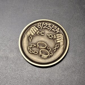 Op de afbeelding: Een ronde, bronskleurige munt met twee handen die een ketting breken, met de datum "8/23/24" bovenaan. Daaronder is een paar handboeien verbonden met een ketting. De munt heeft een gedetailleerd, reliëf ontwerp.