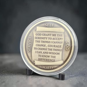 May include: A round bronze-toned medallion encased in a clear plastic protective shell. The center features a scroll design with the text: GOD GRANT ME THE SERENITY TO ACCEPT THE THINGS I CANNOT CHANGE, COURAGE TO CHANGE THE THINGS I CAN, AND WISDOM TO KNOW THE DIFFERENCE. The words SELF, SERVICE, GOD, and SOCIETY are embossed around the perimeter.