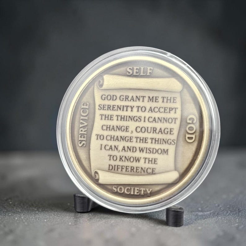 May include: A round bronze-toned medallion encased in a clear plastic protective shell. The center features a scroll design with the text: GOD GRANT ME THE SERENITY TO ACCEPT THE THINGS I CANNOT CHANGE, COURAGE TO CHANGE THE THINGS I CAN, AND WISDOM TO KNOW THE DIFFERENCE. The words SELF, SERVICE, GOD, and SOCIETY are embossed around the perimeter.