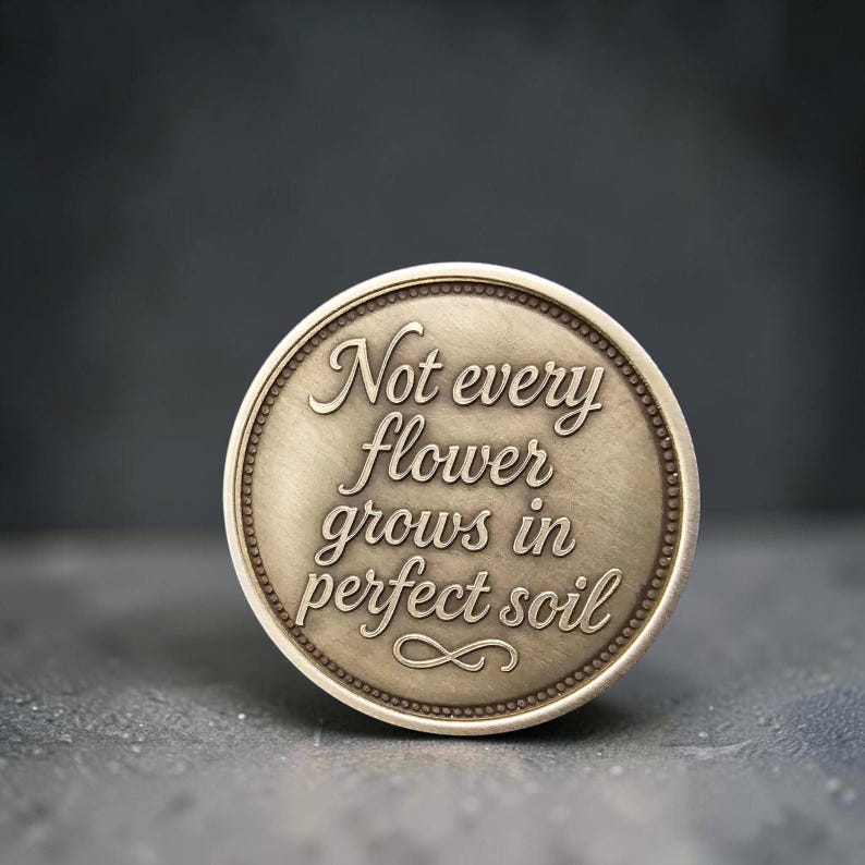 May include: A circular brass-toned metal token featuring a beaded border and the engraved text "Not every flower grows in perfect soil" in a cursive script, with a small decorative flourish centered at the bottom of the coin-like object.