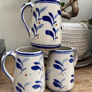 Peut inclure: Trois mugs en céramique blanche avec des motifs floraux bleus. Chaque mug a une anse et est décoré de feuilles bleues et de petits points orange et verts. Les mugs sont empilés sur une surface en bois.