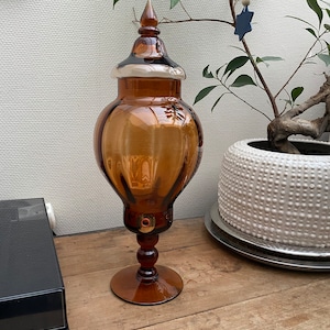 May include: A large amber glass jar with a lid and a pedestal base. The jar has a textured surface and a small hole in the side. The jar is sitting on a wooden table.