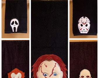 May include: Black hand towels with embroidered horror movie character faces. The characters include Ghostface, Jason Voorhees, Pennywise, Chucky, and Jigsaw. The towels are hanging on a white bar against a tan wall.