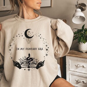 May include: A beige sweatshirt with a black graphic design featuring a crescent moon, stars, a snake, a book, and the text "In My Fantasy Era".