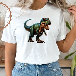 May include: A white t-shirt with a green and blue dinosaur graphic. The dinosaur is facing forward with its mouth open and has a fierce expression.