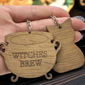 May include: Wooden keychain with two charms: a cauldron with the text "Witches Brew" and a black cat. The charms are brown with a wood grain texture.