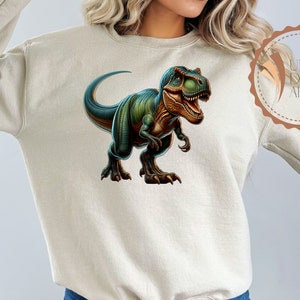 May include: A beige sweatshirt with a green and brown dinosaur graphic on the front. The dinosaur is a Tyrannosaurus Rex with its mouth open and its teeth showing. The dinosaur is facing to the right.