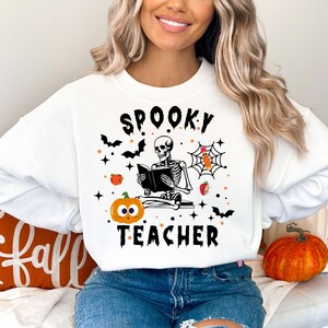 Könnte beinhalten: Weißer Pullover mit einer schwarzen Grafik eines Skeletts, das ein Buch liest, mit dem Text "Spooky Teacher" und Halloween-Motiven.