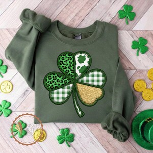 May include: Green sweatshirt featuring a large shamrock design. The shamrock is divided into sections with leopard print, cow print, white and green gingham, and gold glitter. Additional shamrock and gold coin decorations are scattered around the sweatshirt.