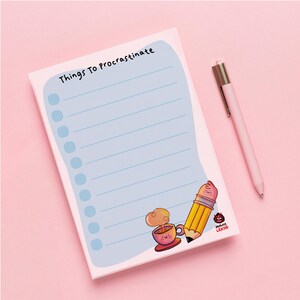 May include: A notepad with the text "Things To Procrastinate" in black, featuring a light blue background with lines and blue circles. A pink pen rests beside the notepad. The notepad has a cartoon illustration of a pencil and a cup of coffee.