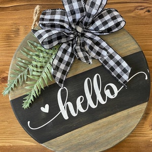 Puede incluir: Un letrero redondo de madera con un lazo de cinta a cuadros blanco y negro, follaje verde y la palabra "hello" escrita en cursiva blanca con un pequeño corazón.
