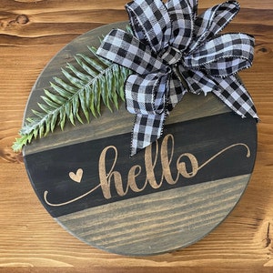 Op de afbeelding: Een rond houten bord met een zwart-wit geblokte strik, een groene varen en het woord "hello" in gouden letters. Het bord heeft een zwart-grijze gestreepte achtergrond.