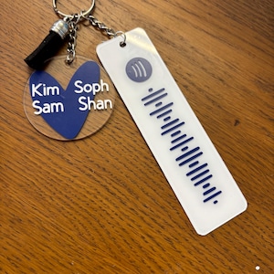 May include: A personalised keyring with a clear acrylic charm featuring names and a blue heart. A rectangular tag with a blue music code and Spotify logo is attached. The keyring has a silver chain and a black tassel.