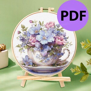 以下が含まれることがあります： 紫、ピンク、青の花でいっぱいのティーカップのクロスステッチ刺繍。ティーカップはソーサーの上にあり、刺繍は木製フープで額装されています。