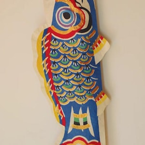 Könnte beinhalten: Ein farbenfroher, fischförmiger Koinobori-Windsack, der traditionell in Japan zum Kindertag verwendet wird. Der blaue Fisch hat komplizierte Designs in Rot, Gelb und Grün, mit einem großen Auge und Schuppenmustern. Er ist an einer roten Schnur aufgehängt.