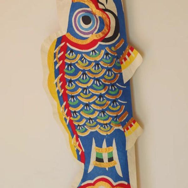 Puede incluir: Un Koinobori en forma de pez, tradicionalmente utilizado en Japón para celebrar el Día del Niño. El pez azul presenta intrincados diseños en rojo, amarillo y verde, con un ojo grande y patrones de escamas. Está suspendido por una cuerda roja.