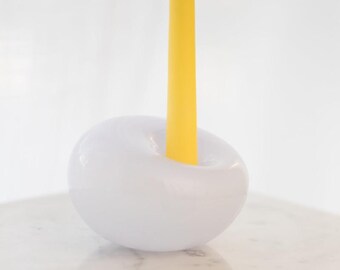 May include: A smooth, white, doughnut-shaped candle holder with a glossy finish, featuring a single, tall, bright yellow taper candle inserted into the central opening, creating a stark contrast between the white base and the vibrant yellow wax.