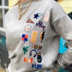 Könnte beinhalten: Ein hellgrauer Pullover mit verschiedenen bunten gestickten Aufnähern, darunter ein Würfel, ein Kleeblatt, ein Blitz, ein Stern, eine Flasche, ein Kopfhörer, ein Boxhandschuh und Wörter wie "Hey", "Oh La La", "Love", "Sssst" und "Fun".