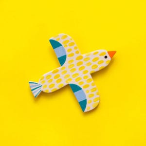 Könnte beinhalten: Ein gelber Holzvogel mit blauen und weißen Punkten und einem orangefarbenen Schnabel. Der Vogel fliegt mit ausgebreiteten Flügeln.