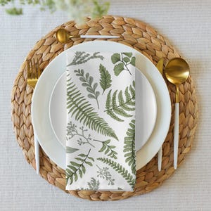 Puede incluir: Una servilleta de tela blanca con un patrón de helechos y hojas verdes. La servilleta está doblada y colocada sobre un plato blanco con un borde dorado. El plato está sobre un mantel individual de mimbre tejido.