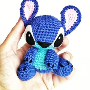 May include: A blue and turquoise crocheted Stitch plush toy with pink ears and black eyes. The toy is sitting in a person's hand.